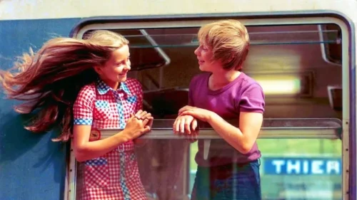 Due giovani protagonisti del film Gli anni in tasca si guardano e sorridono mentre sono affacciati al finestrino di un treno in corsa